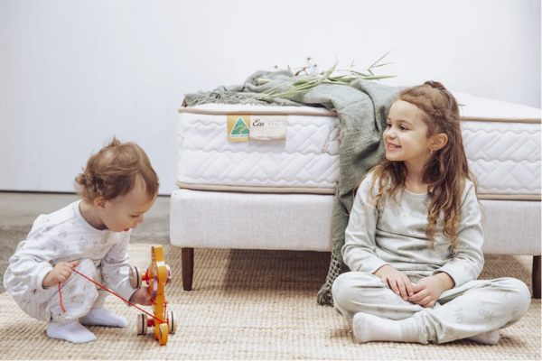Two children playing with a toy in front of a mattress with an 'Eco Kids' brand label.