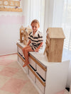 Child sitting on a wooden Boori Kids Tidy Toy Cabinet pctured in the Barley White & Almond Colour in a bright room with white curtains.